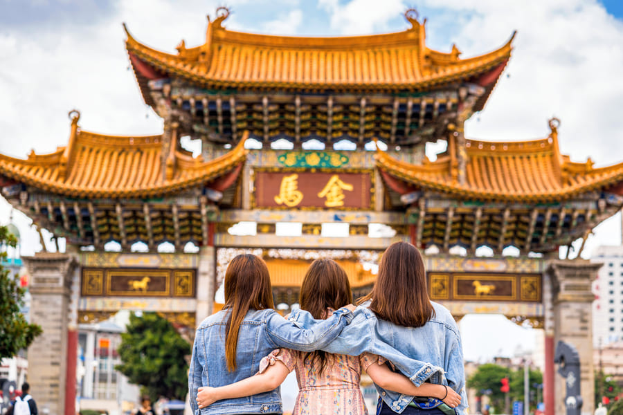 Tres jóvenes abrazadas de espaldas frente a una impresionante puerta tradicional china con techos dorados y ornamentación clásica.